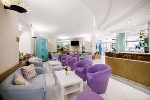 A modern lobby with purple and blue seating, a reception desk, and a TV mounted on the wall. - Hotel Ariane (Rimini)