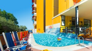 Uma área de piscina com cadeiras de sol azuis e vermelhas, um brinquedo flutuante amarelo e uma piscina azul cristalina.