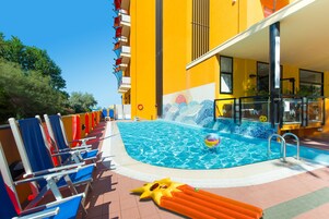 Un área de piscina con sillas de descanso azules y rojas, un flotador inflable amarillo y una piscina azul cristalina.