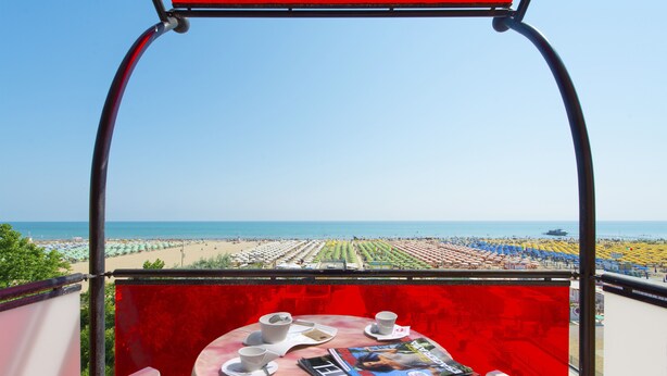 Uma mesa com xícaras e uma revista, com vista para uma praia com fileiras de guarda-sóis e cadeiras.
