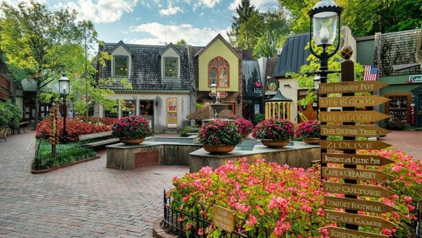 A quaint village square with cobblestone paths, flower beds, and a directional signpost.