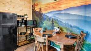 A compact kitchen with a refrigerator, microwave, and coffee maker, a wooden dining table with chairs, and a scenic mural on the wall.