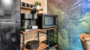 A kitchenette with a refrigerator, coffee maker, microwave, and a shelf with mugs and a small plant.