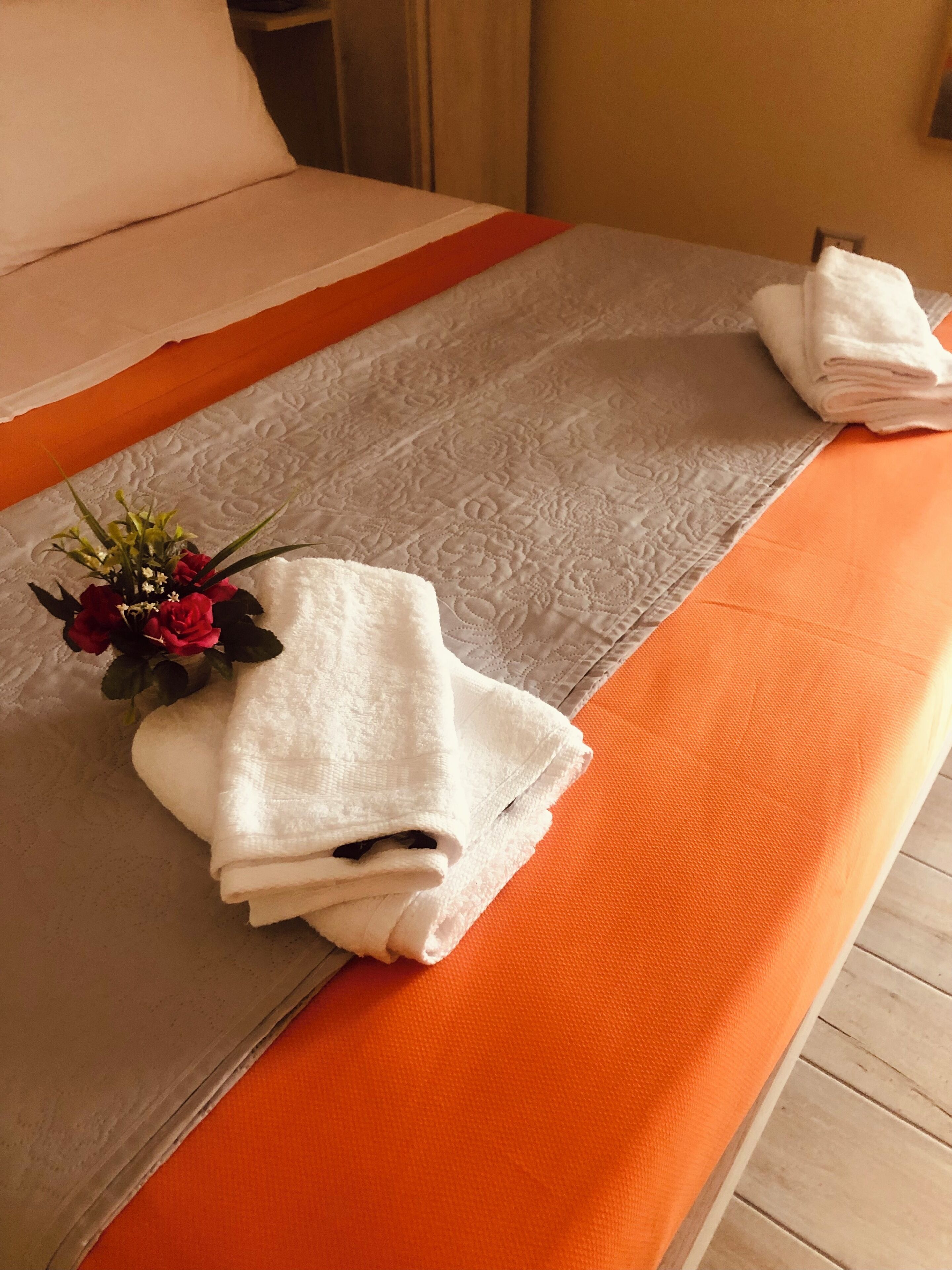 A neatly made bed with a white and orange bedspread, a folded white towel, and a small bouquet of flowers.