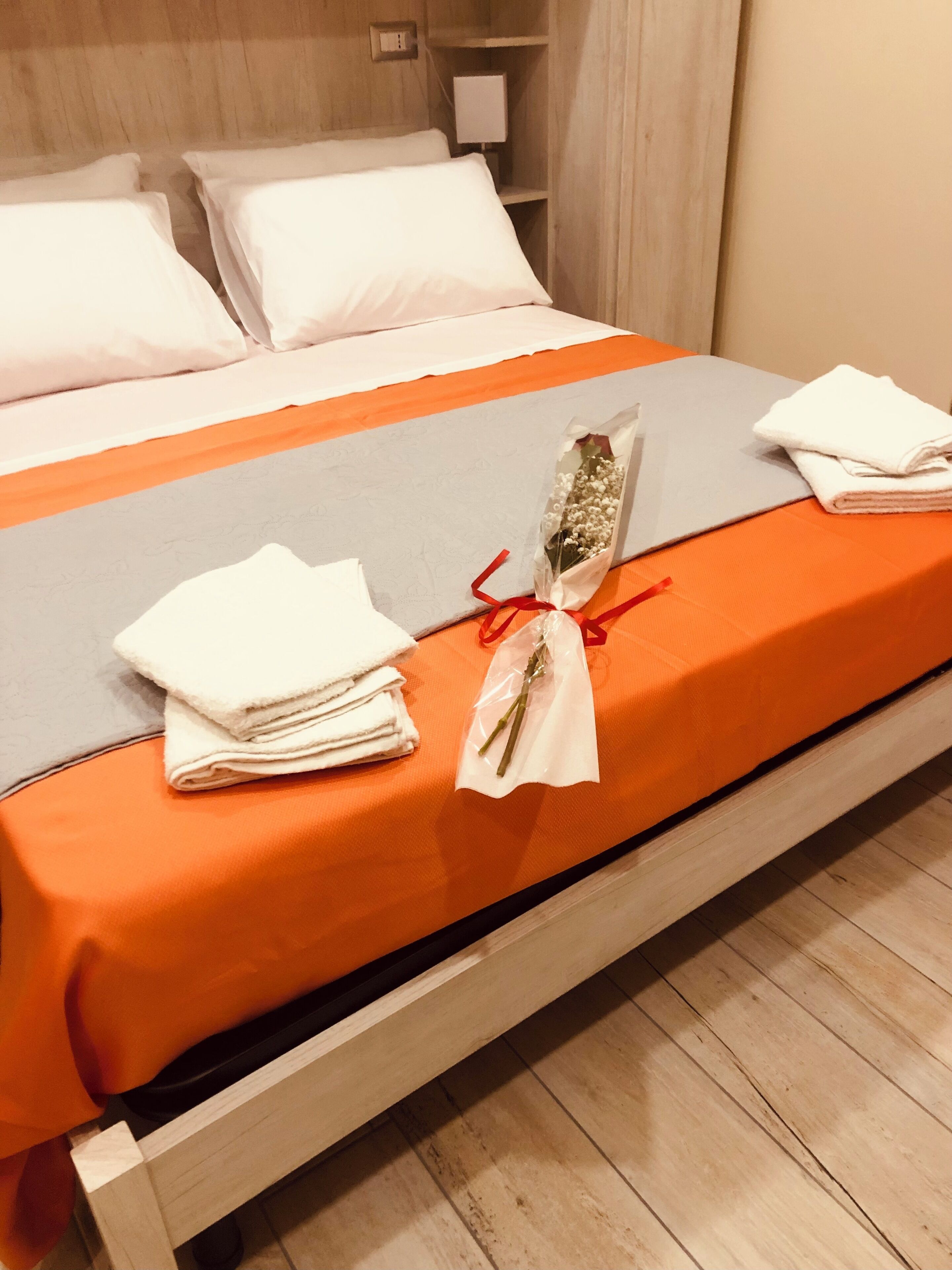 A neatly made bed with white and orange bedding, a small bouquet of flowers, and folded towels.