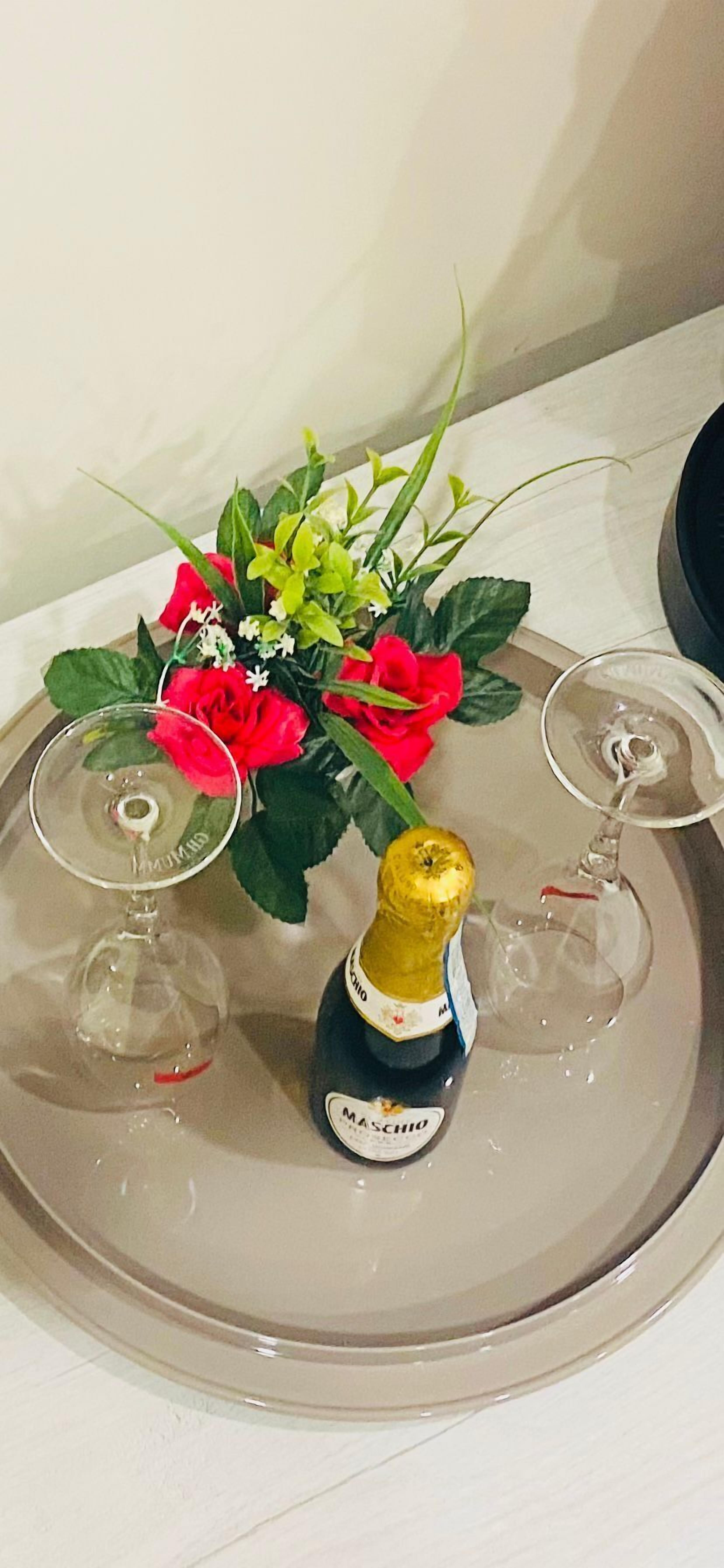 Une table sur laquelle se trouve un plateau en verre, avec une bouteille de champagne Mascaraio, un verre et un bouquet de fleurs rouges.