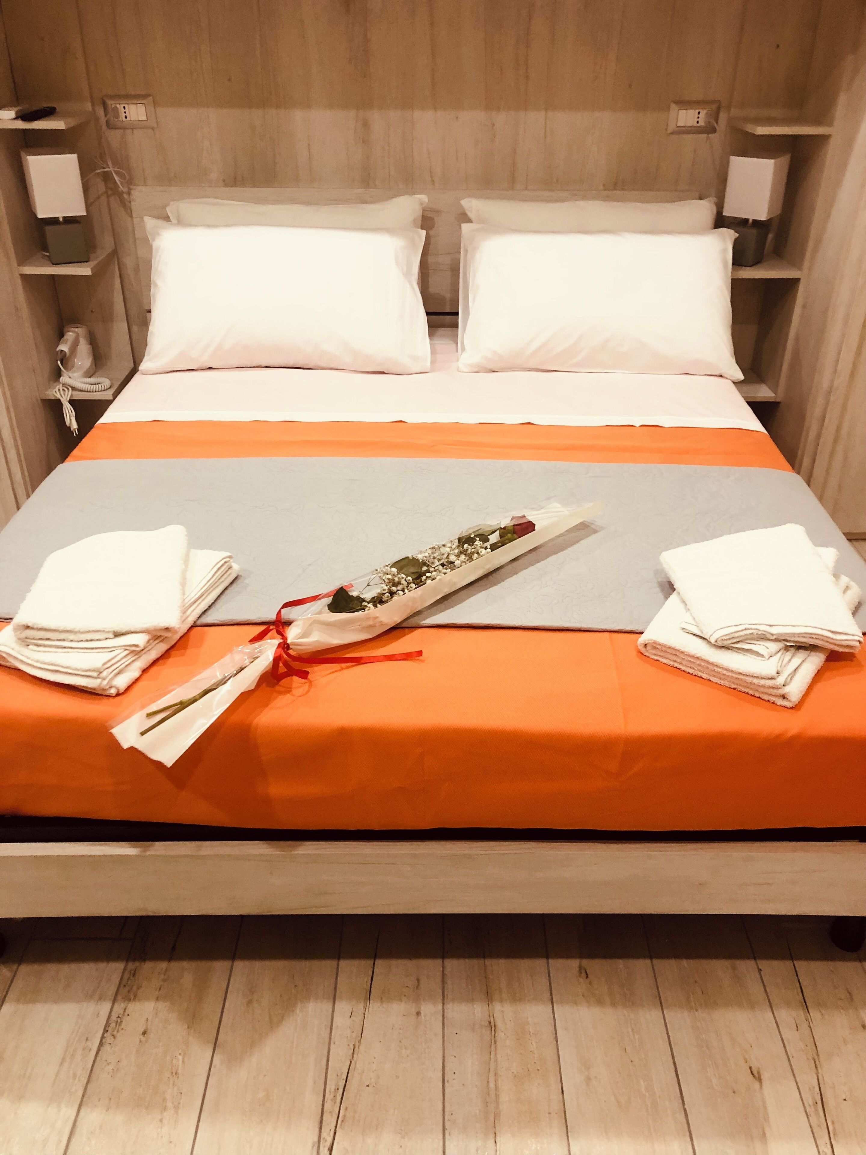 A neatly made bed with white pillows and an orange bedspread, two folded white towels, and a small bouquet of flowers tied with a red ribbon on the bed.