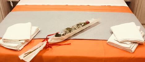A neatly made bed with white pillows and an orange bedspread, two folded white towels, and a small bouquet of flowers tied with a red ribbon on the bed.