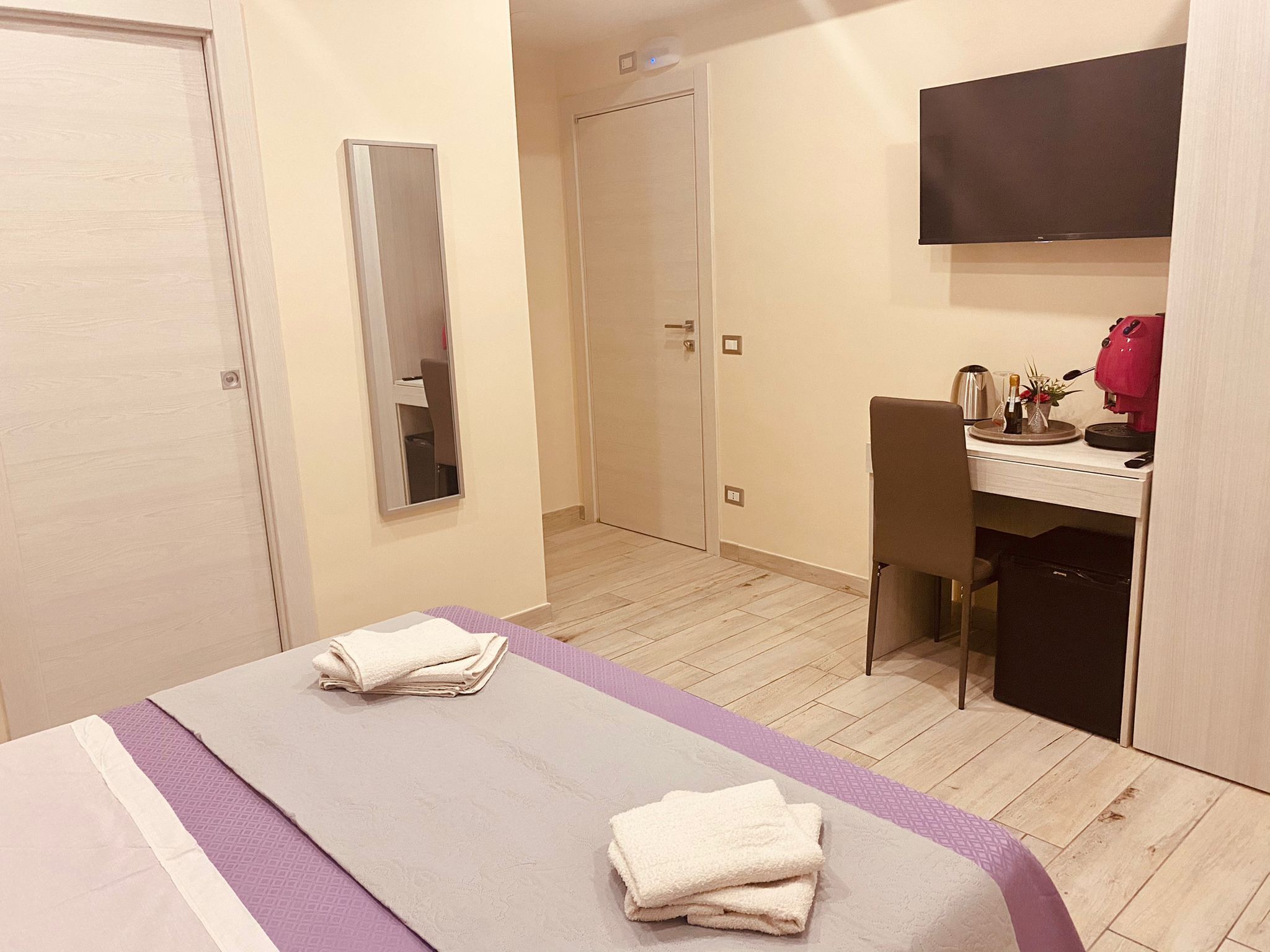 A hotel room with a bed, a desk, a chair, a mirror, and a TV.