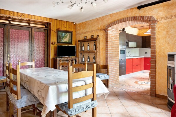 A dining area with a table set for four, wooden chairs, a television, and a brick oven in the background.
