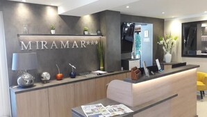 A reception desk with a modern design, featuring a nameplate that reads "MIRAMAR" and a reception area with a counter, seating, and a vase with flowers. - Hôtel Miramar Cap d'Antibes (Antibes)