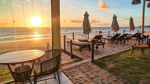 Un patio frente a la playa con vista al atardecer sobre el océano, mesas y sillas de madera, y sombrillas de playa.