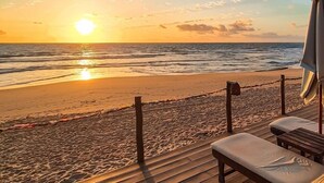 Una terraza de madera con una silla de descanso y una sombrilla, con vista a una playa donde se pone el sol sobre el océano.