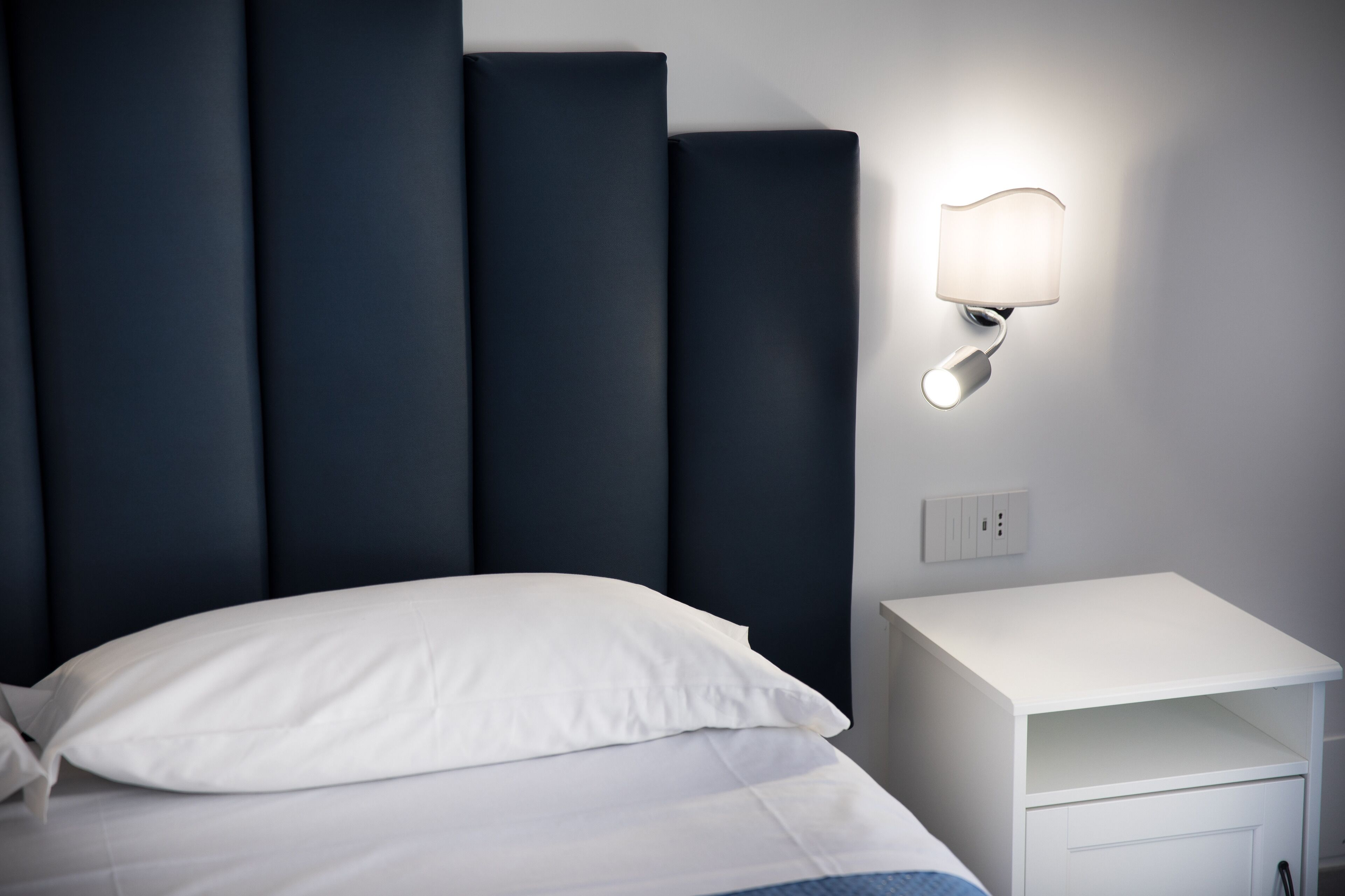 A neatly made bed with white bedding and a single white pillow, a bedside table with a lamp, and a wall socket.