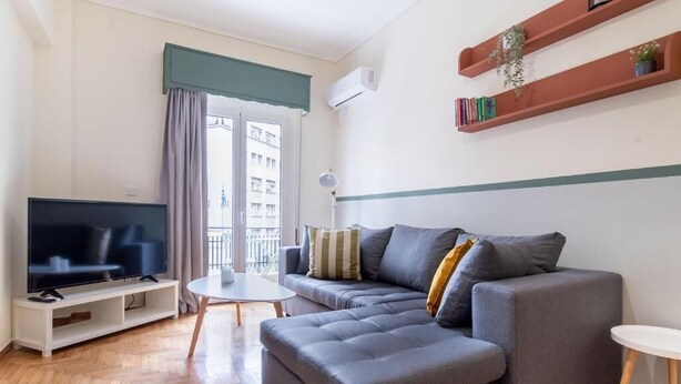 Een moderne woonkamer met een grijze modulaire bank, een flatscreen televisie op een witte standaard, een klein rond tafeltje en een boekenkast met boeken en planten.