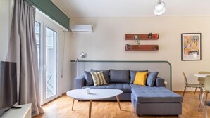 Een moderne woonkamer met een grijze modulaire bank, een witte salontafel en een televisie op een witte standaard. Er is een raam met gordijnen, een wandairconditioner en een lijstwerk kunstwerk aan de muur.