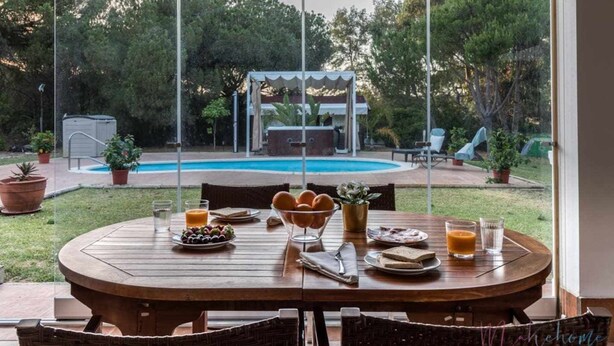 Un tavolo da pranzo in legno con oggetti per la colazione, una piscina e un'area salotto coperta in un giardino.