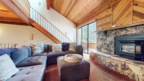 A living room with a stone fireplace, a wooden staircase, and a large window. - Pet-Friendly Rustic Home, Washer/Dryer, Mountain Views, Steps to the Shuttle (Mammoth Lakes)