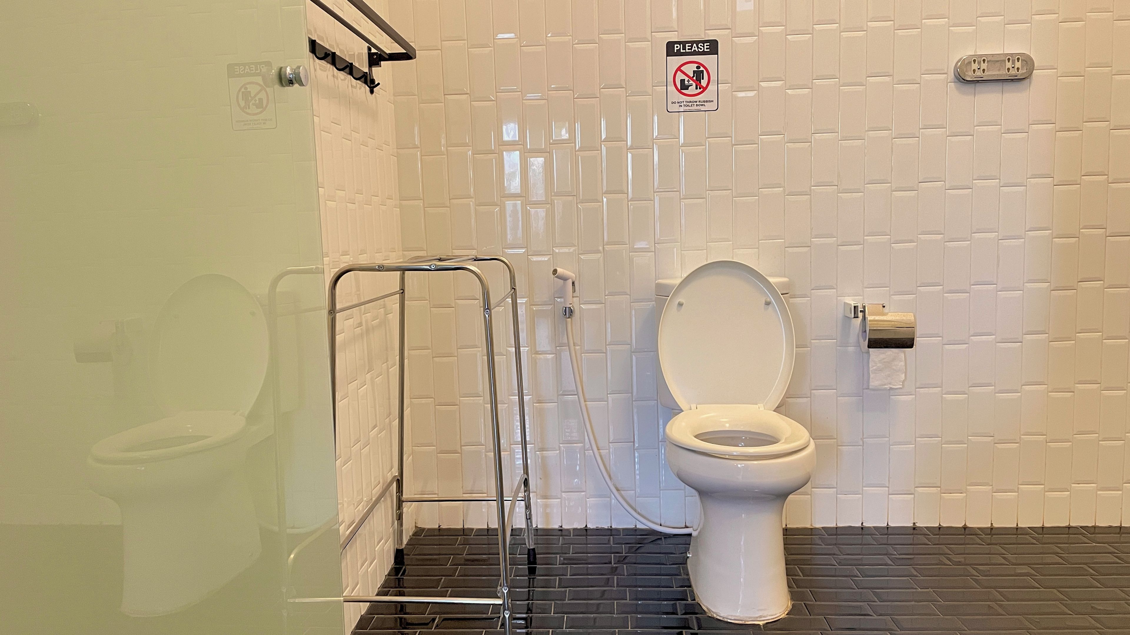 Un baño con inodoro blanco, barandilla, letrero de prohibición de basura y pared con azulejos.