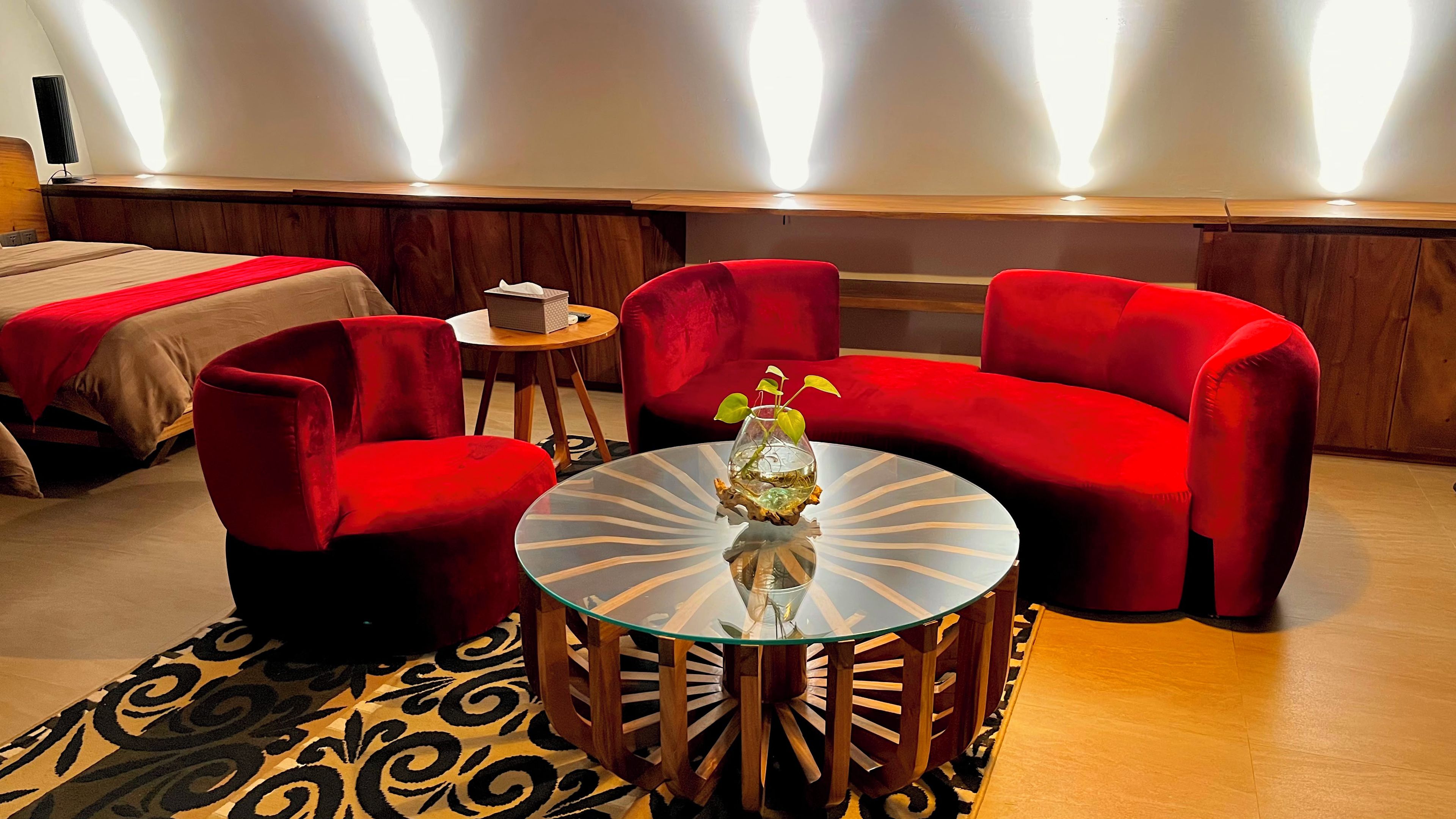 A hotel room with a bed, a red sofa, a red armchair, a glass coffee table with a vase, and a small table with a book.