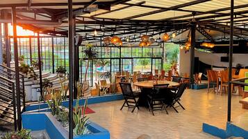 An indoor dining area with tables and chairs, large windows, and hanging lights.