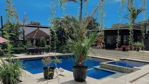 Una piscina rodeada de plantas en macetas, con mobiliario exterior y un edificio al fondo.