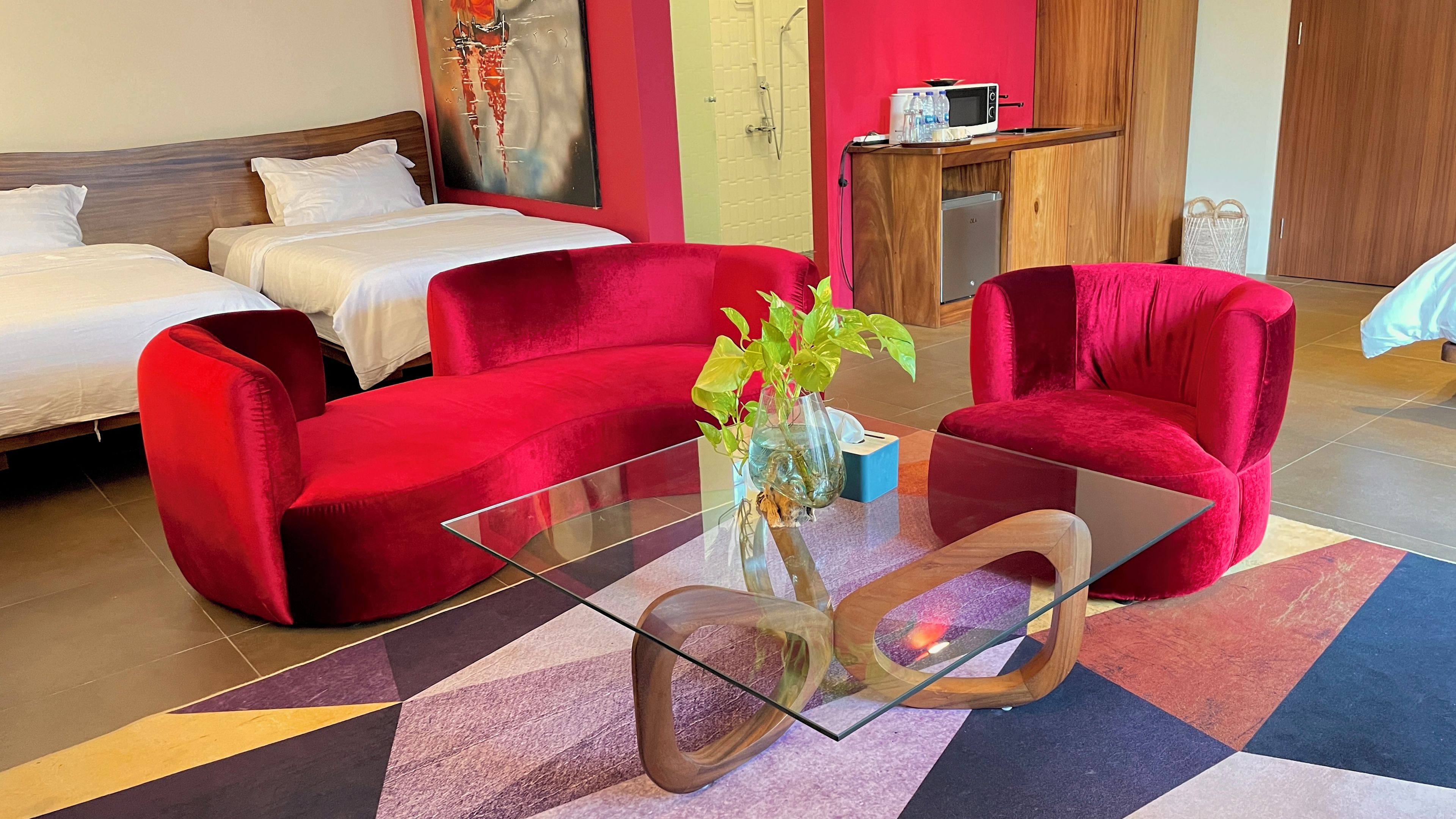 Une chambre d’hôtel avec un lit, deux fauteuils rouges, une table basse en verre et une petite plante.
