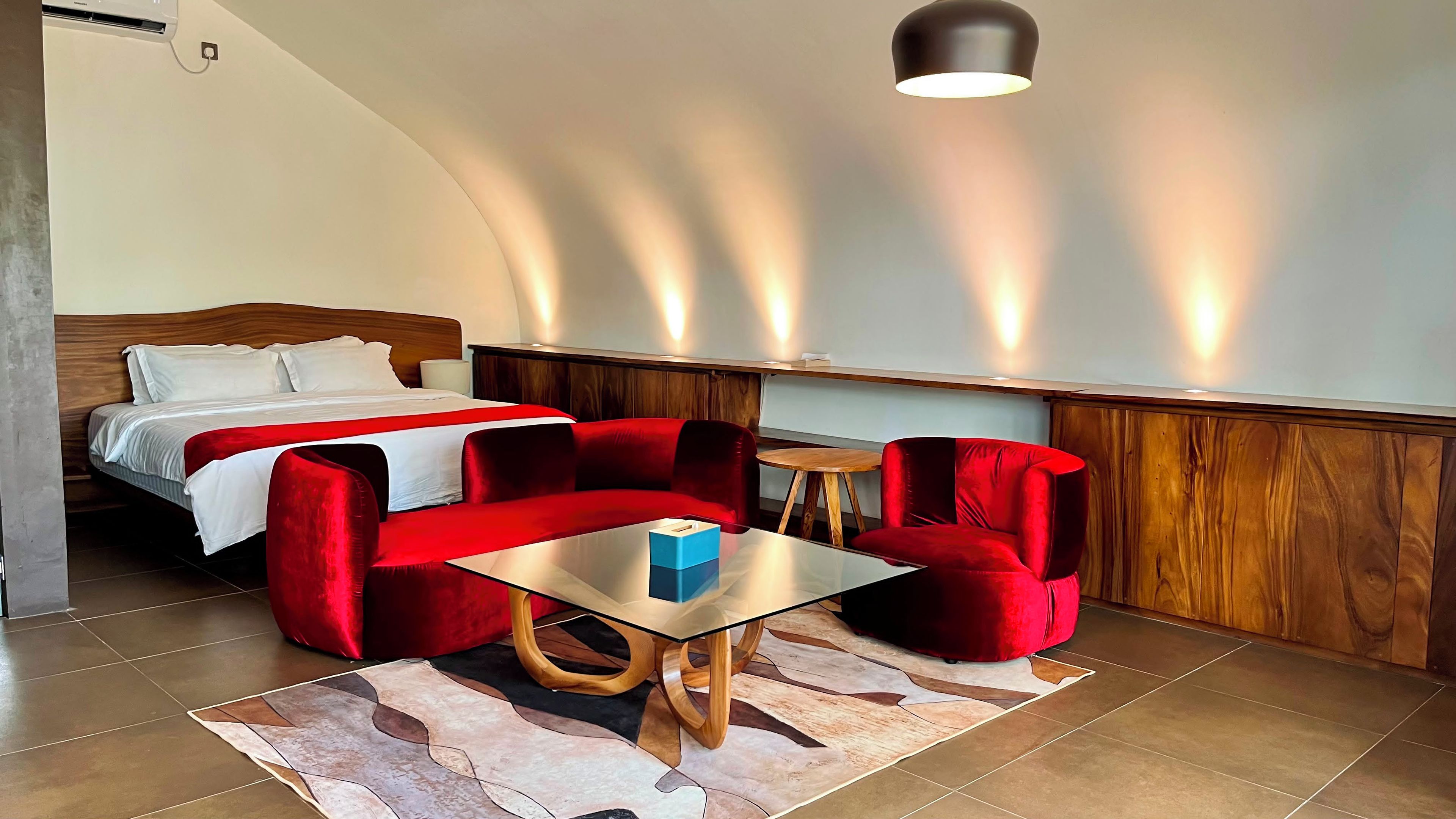 A hotel room with a bed, red upholstered chairs, a glass coffee table, and a wooden side table.