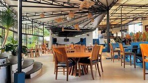 A restaurant with wooden tables and chairs, a high ceiling with hanging lights, and large windows allowing natural light. - Lucca Resort & Residence (Bandengan)