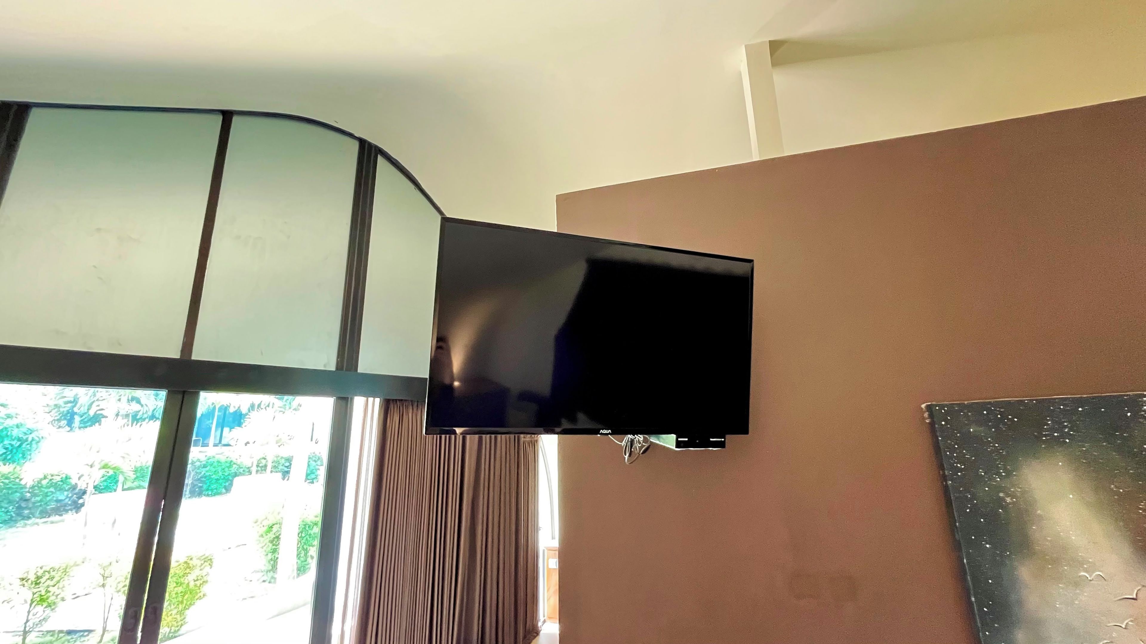 A flat-screen TV mounted on a wall, a window with curtains, and a partial view of a room with a brown wall.