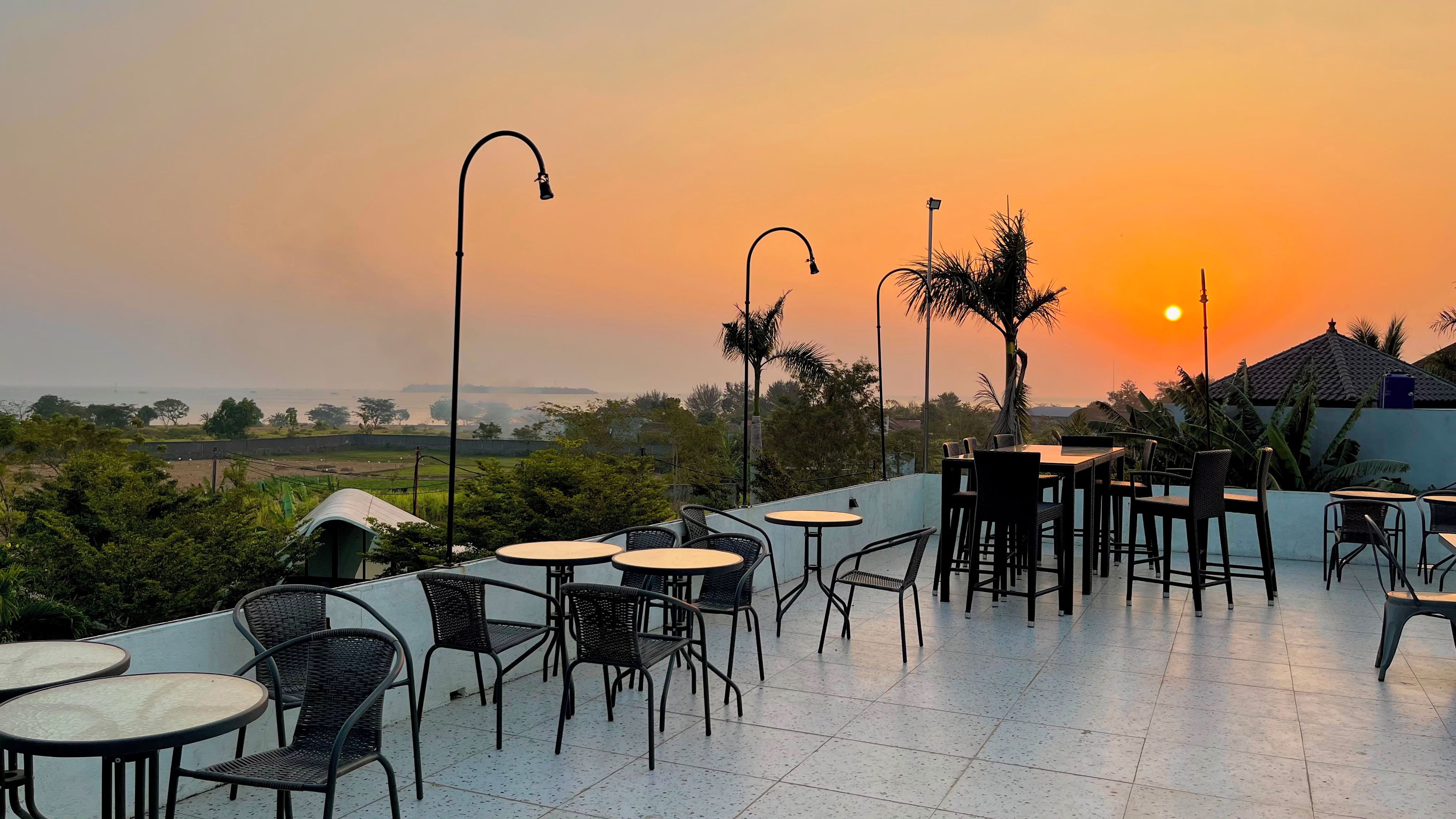 An outdoor terrace with round tables and chairs, street lamps, and a view of the sunset over a landscape with trees and buildings.