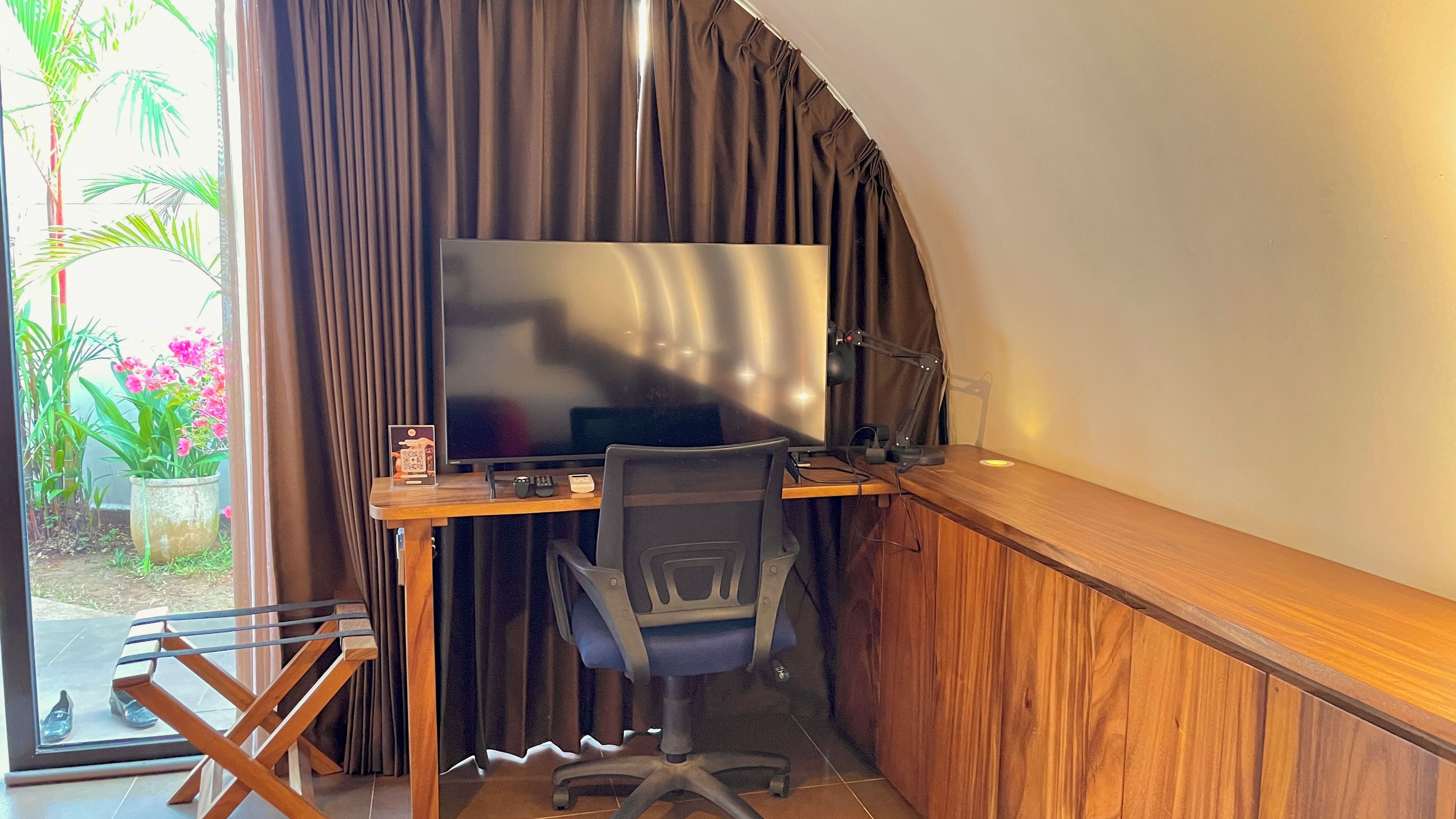Ein Zimmer mit einem großen Flachbildfernseher, einem Holztisch, einem schwarzen Bürostuhl und einem Fenster mit Vorhängen.