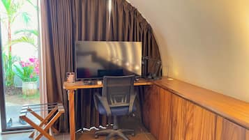 Ein Zimmer mit einem großen Flachbildfernseher, einem Holztisch, einem schwarzen Bürostuhl und einem Fenster mit Vorhängen.