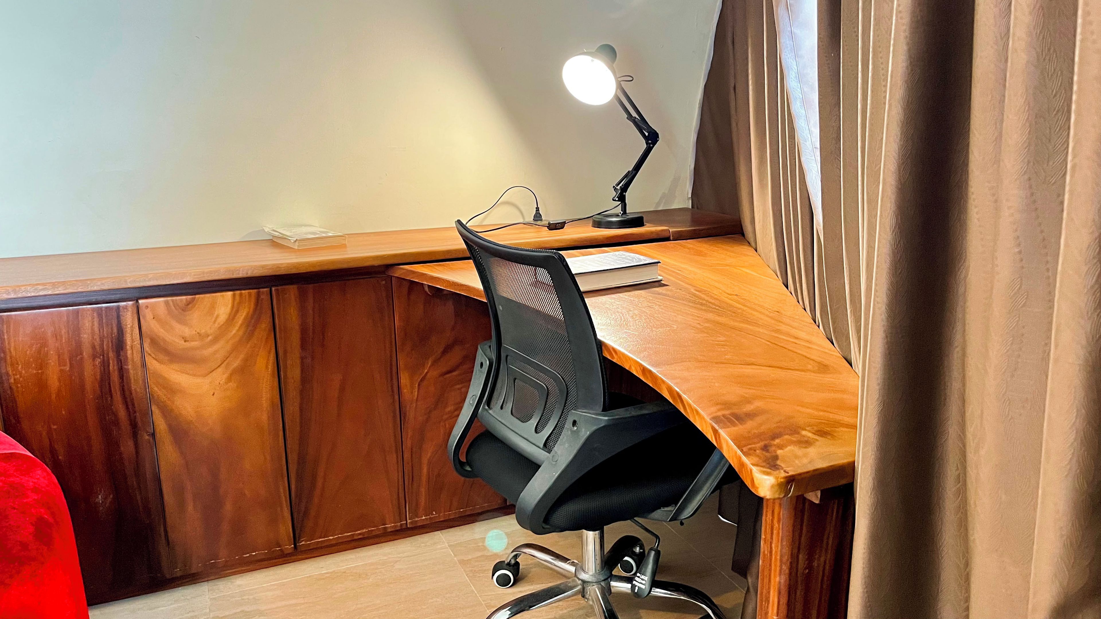 A wooden desk with a black office chair, a desk lamp, and a book.