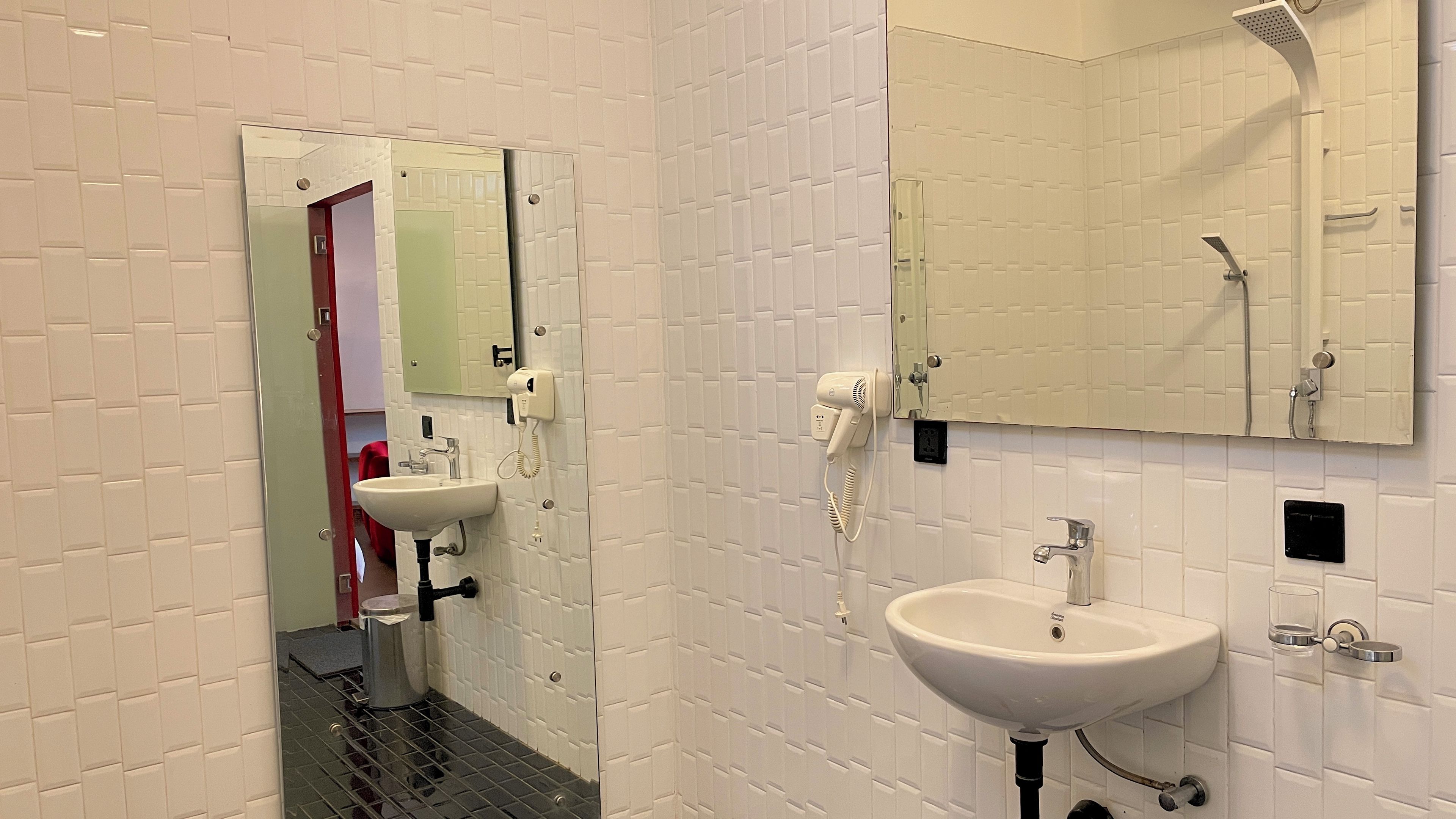 Une salle de bain moderne avec un mur recouvert de carreaux blancs, un lavabo, un miroir, un sèche-cheveux et une pomme de douche.