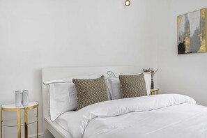 A neatly made bed with white bedding, a small bedside table with a vase, and a wall-mounted abstract painting.