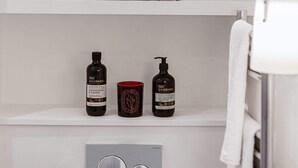 Ein Badezimmer mit einem weißen Regal, das eine Vase mit gelben Blumen, ein gerahmtes Schild mit Herzchen und ein Handtuch beherbergt. Darunter befinden sich zwei Produkte der Marke Goodness sowie ein Spiegel.