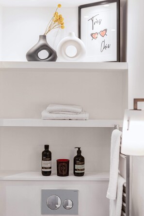 A bathroom with a white shelf holding a vase with yellow flowers, a framed sign with hearts, and a towel. Below, there are two bottles of Goodness brand products and a mirror. - Lady on the Lane, 2 bed Primrose Hill - by Out of Office Lifestyle (London)