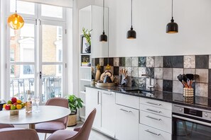 A modern kitchen with white cabinets, a black countertop, and a backsplash with various tiles. There are hanging pendant lights, a round dining table with chairs, and a window with a view of buildings outside.