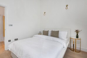Ein Schlafzimmer mit einem weißen Bett, zwei Nachttischen, einer Wandleuchte und einer kleinen Vase mit Blumen auf der rechten Seite.