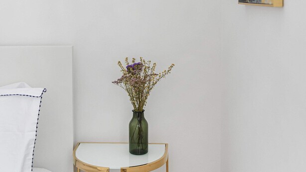 Ein Zimmer mit weißer Wand, einem runden Tisch mit einer grünen Blumenvase und einem Gemälde mit goldenem und grauem abstrakten Design.
