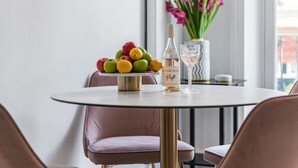 Ein runder Tisch mit Obstschale, einer Flasche und einem Glas. Rosa Stühle stehen um den Tisch herum.