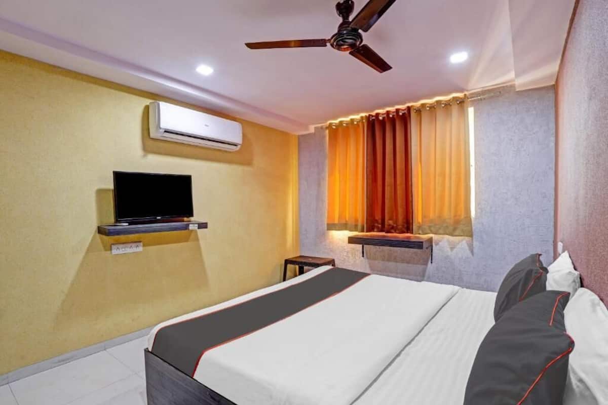 Una habitación de hotel con cama, televisor en un estante, aire acondicionado y ventana con cortinas.