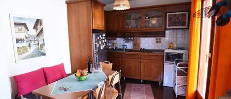 A compact kitchen with wooden cabinets, a dining table with chairs, a refrigerator, an oven, and a hanging light fixture.
