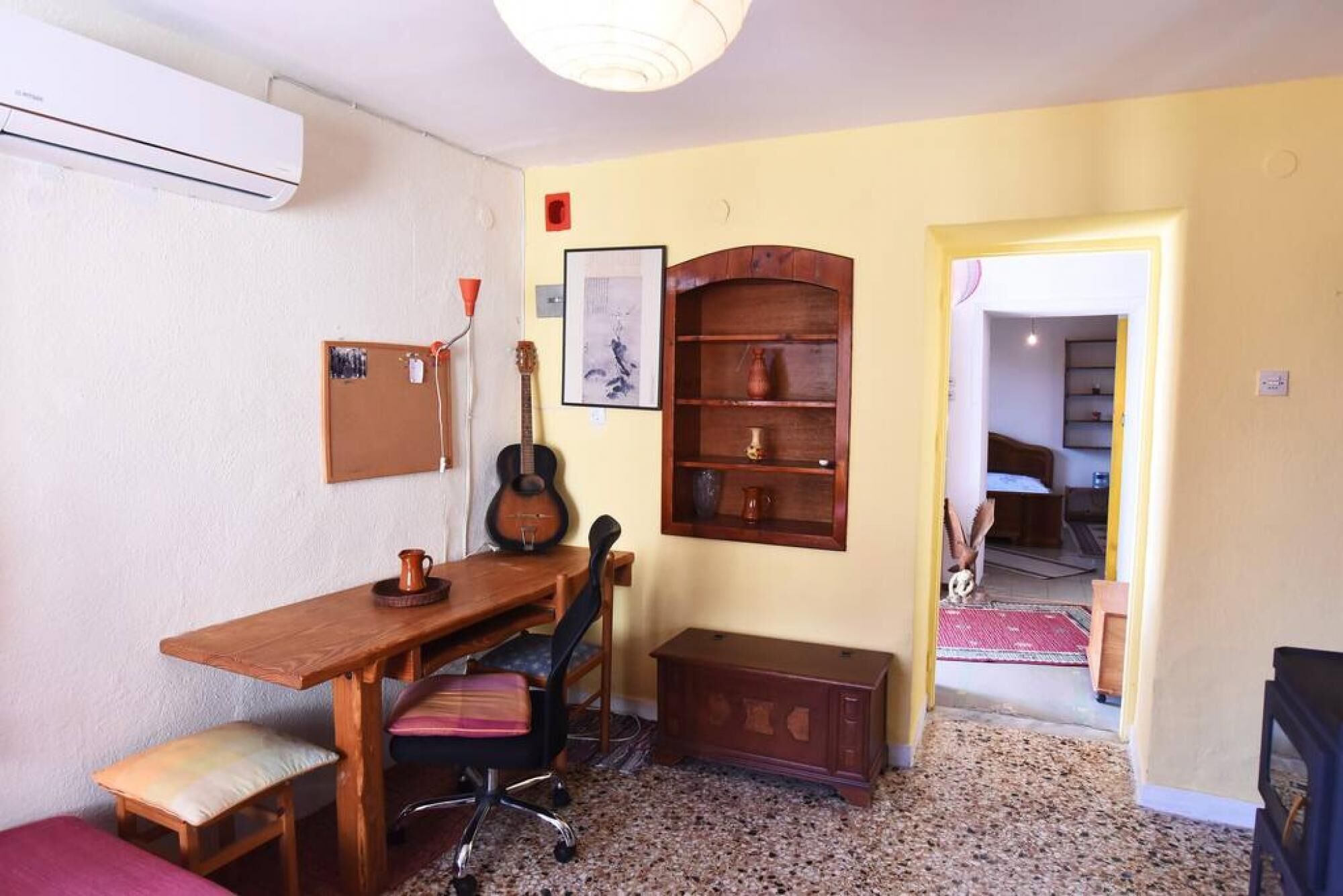 Ein kleines Zimmer mit einem Holztisch, einem Stuhl, einer Gitarre, einer Pinnwand und einem Bücherregal.
