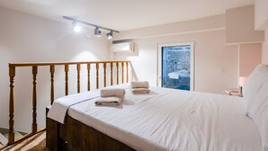 Un dormitorio con estructura de cama de madera, ropa de cama blanca, una mesita de noche con lámpara y una ventana con vista a árboles.