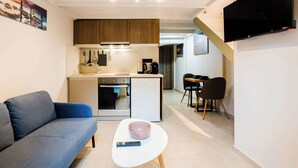 Una sala de estar moderna con un sofá, una mesa de centro blanca, un control remoto, una zona de cocina con un refrigerador, estufa y microondas, y un televisor montado en la pared.