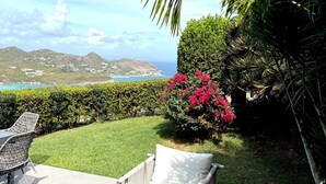 A patio with outdoor furniture, a view of the ocean, and a landscaped garden with a bush and palm tree.