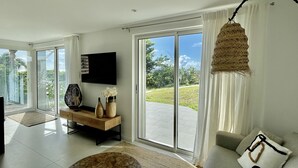 A modern living room with a large TV, a sofa, a coffee table, and a view of a garden through sliding glass doors.