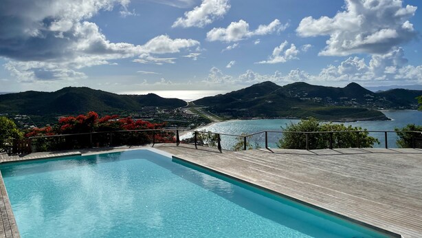 A clear blue swimming pool with a wooden deck, surrounded by lush greenery and red flowers. In the background, there are mountains and a view of the sea.